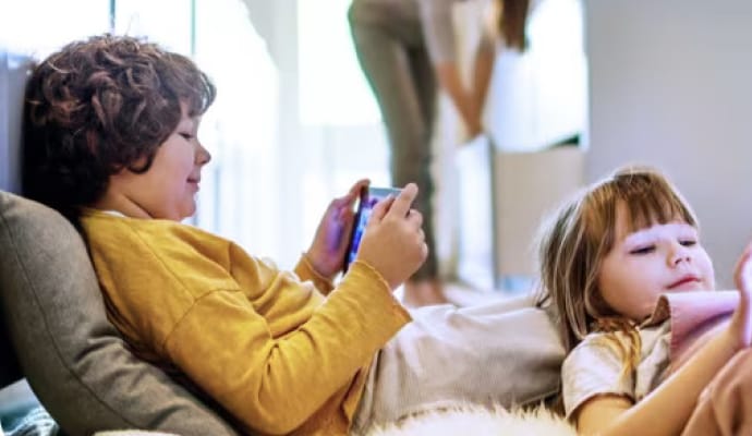 Kids lying down using tablet devices while mother in the background is tidying the kitchen.